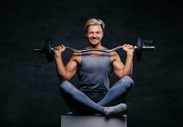 Positive and smiling blond sporty male is dressed in a grey sportswear sits on a box and holds barbel.