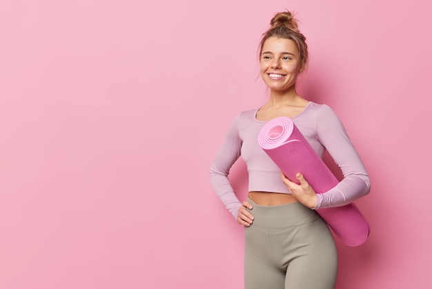 Positive smiling slim woman in tracksuit poses with rolled karemat does yoga pilates or fitness exercises has healty body isolated over pink background blank space aside for your promotional content