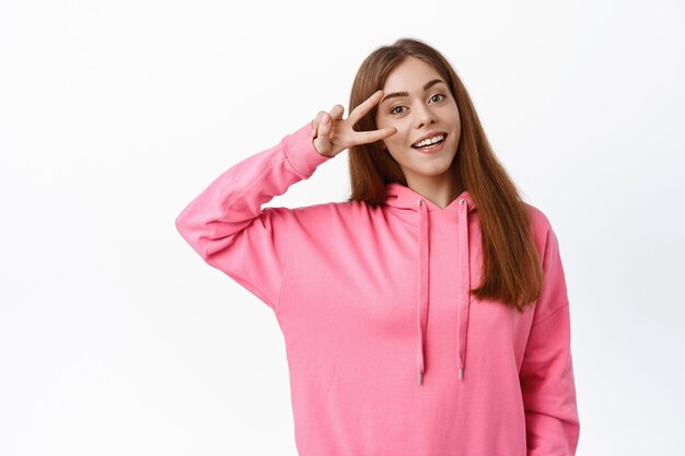 Positive smiling woman shows peace sign and looks happy, making v-sign gesture, express positive and carefree emotions, white wall