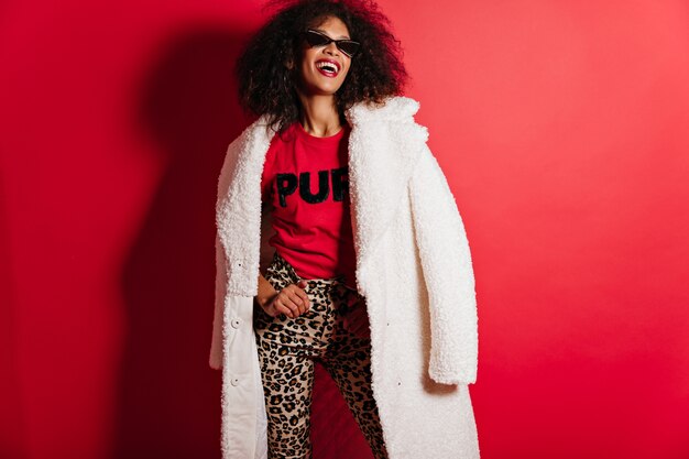 Positive smiling woman in sunglasses standing on red wall