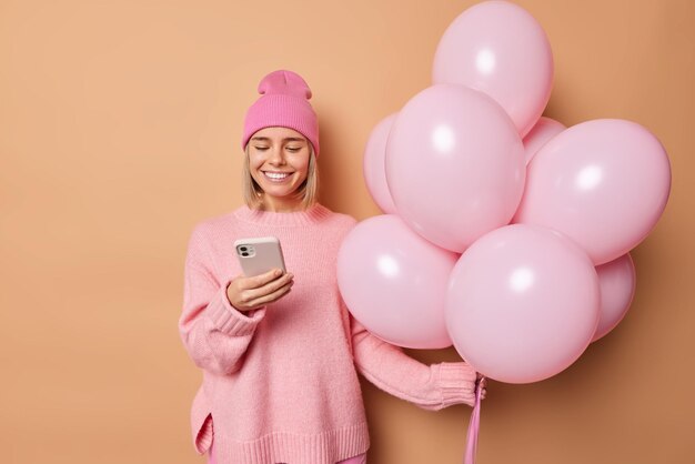 긍정적인 미소를 짓고 있는 여성은 모자를 쓰고 점퍼는 스마트폰의 메시지 내용을 확인합니다. 생일 축하를 위해 많은 풍선을 들고 베이지색 스튜디오 벽에 격리된 파티를 조직합니다