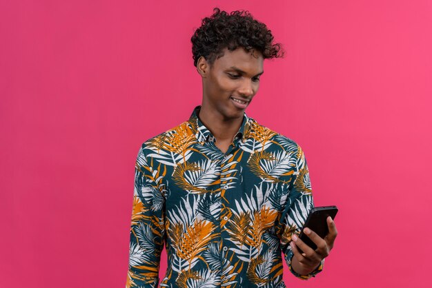 A positive and smiling young handsome dark-skinned man with curly hair in leaves printed shirt looking at his mobile phone 