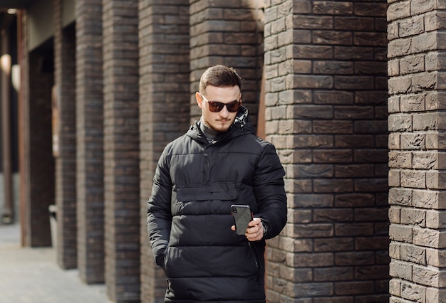 Positive smiling young male in stylish clothes standing outside alone near wall of urban building and speaking on mobile phone