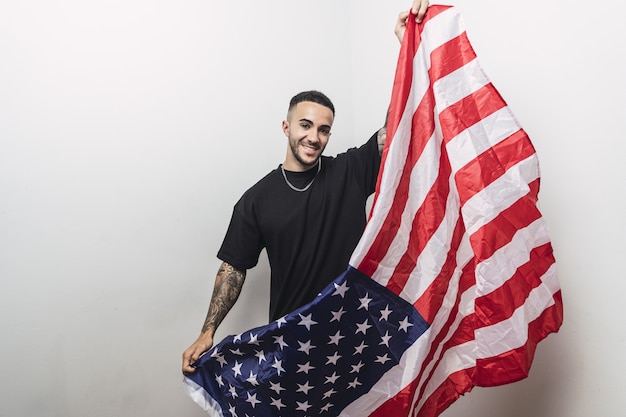Позитивный испанский мужчина с татуированными руками позирует с американским флагом, изолированным на белой стене