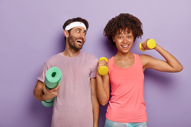 Positive sportsman wears headband and t shirt, holds crumpled fitness mat, looks gladfully at girlfriend who raises arms with dumbbells, have workout together