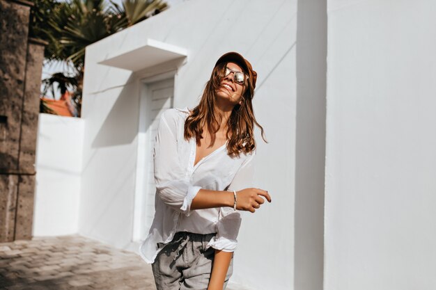 Positive stylish woman in gray pants and oversized shirt having fun on street next to white building and palm trees.