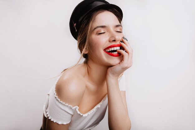 Positive stylish woman with bright lipstick laughing with her eyes closed. Girl in black cap posing on white wall.