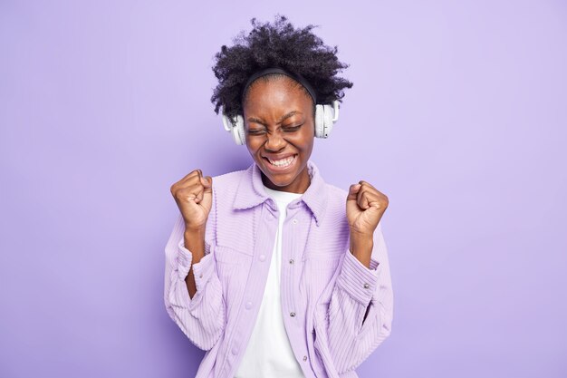 Positive successful Afro American woman clenches fists from joy listens to  new music playlist via wireless headphones