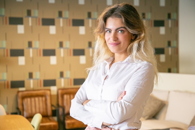 Free photo positive successful businesswoman posing with arms folded in co-working or coffee shop interior, looking at camera and smiling