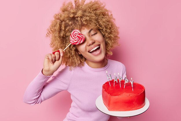 Positive sugar addicted woman covers eye with lollipop smiles happily holds heart shaped cake with candles enjoys eating tasty sweet deserts wears casual jumper isolated over pink background