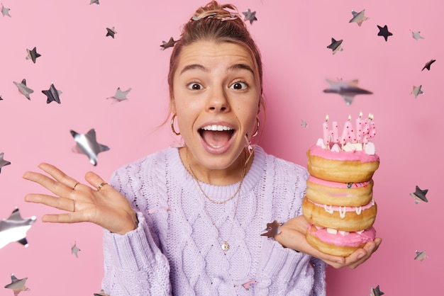 무료 사진 긍정적인 놀란 생일 소녀는 축하를 받고 즐거운 느낌을 받습니다. 축제용 달콤한 도넛은 캐주얼한 점퍼를 입고 주위에 색종이 조각이 있는 분홍색 배경 위에 고립된 축하에 자유 시간을 보냅니다