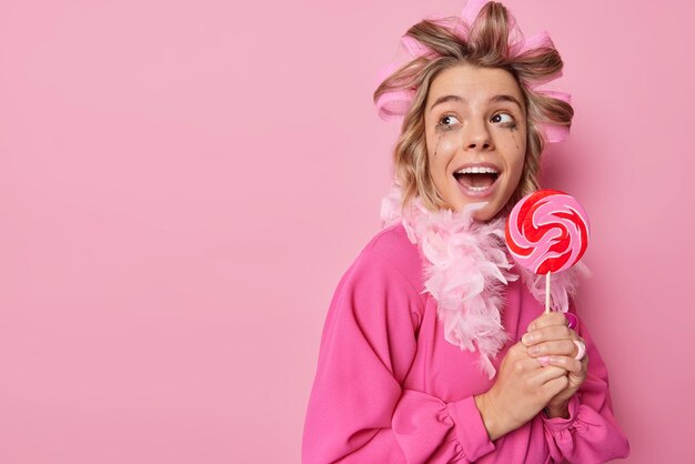 Positive surprised woman looks away with widely opened mouth has sweet tooth holds lollipop on stick applies hair rollers and dress isolated over pink background blank space for your promotion