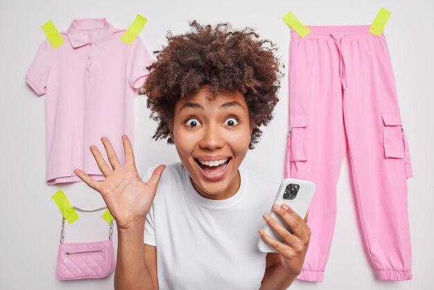 Позитивно удивленная молодая женщина с вьющимися волосами держит ладонь поднятой, радостно смотрит в камеру, использует смартфон для прокрутки интернет-поз на белом фоне, намазанные предметы одежды позади