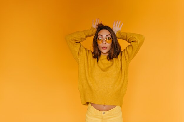 Positive tanned girl in sunglasses making funny faces on orange wall. Indoor portrait of cheerful brown-haired woman wears orange sweater.