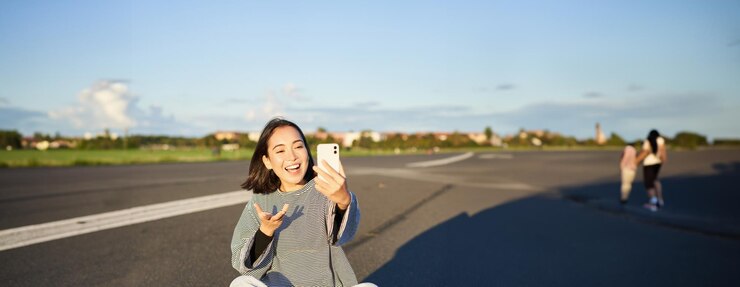 無料Photo ポジティブなティーンエイジャーのアジア人女の子は ⁇ 携帯電話アプリでセルフィービデオチャットをし ⁇ スケートボードに座ってレコードを録音します ⁇ 