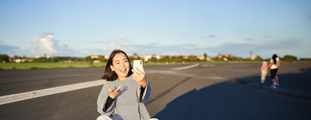 무료 사진 긍정적 인 십대 아시아 소녀는 휴대 전화 앱에서 셀카 비디오 채팅을하고 스케이트보드에 앉아 기록합니다.