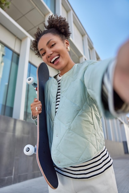 Позитивная девочка-подросток с пучками волос делает селфи на фото, на котором она держит скейтборд, наслаждается любимыми позами для хобби в городских условиях, улыбается и носит уличную одежду.