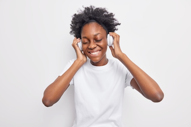 Positive teenage girl with natural curly hair expresses authentic emotions smiles gently keeps eyes closed wears stereo headphones dressed in casual t shirt isolated on white 