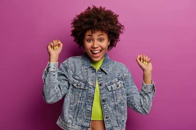 Positive teenage girl with natural curly hair raises arms, clenches fists, has upbeat mood, rejoices success, wears denim jacket, isolated over purple wall, dances to favorite music at party