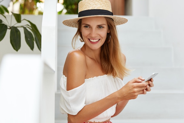 肯定的な思いやりのあるリラックスした女性の夏帽子、ワイヤレスインターネットに接続されている現代の携帯電話でメッセージを入力すると、スタイリッシュな服を着ています。