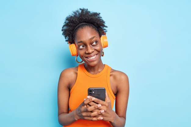 Positive thoughtful woman with natural curly hair dark skin uses mobile phone application listens to music