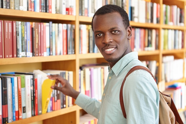 Positive traveler carrying backpack picking book in bookstore