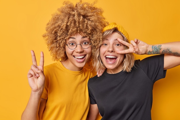 Позитивные две подруги показывают жест мира улыбаются радостно смеются, счастливо одетые в повседневные футболки, изолированные на желтом фоне Игривые девушки сходят с ума, хорошее настроение делает знак победы