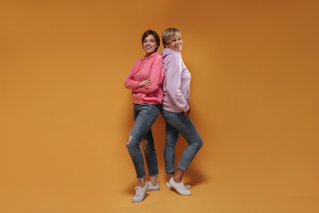 Positive two short haired ladies with lovely smiles in trendy pink sweatshirt, modern jeans and cool sneakers looking into camera on orange backdrop.