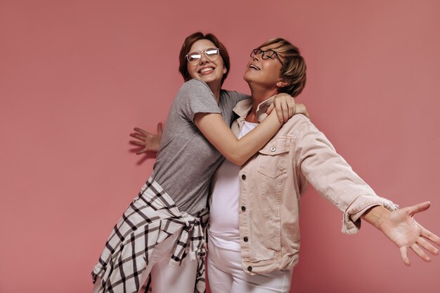 Positive two short haired women with modern glasses in fashionable light clothes hugging and smiling on pink background.