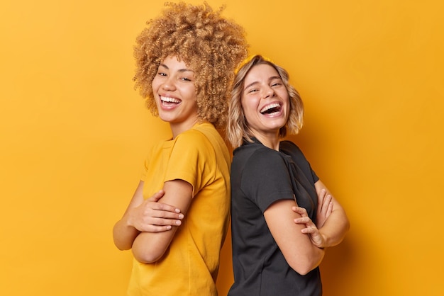 Positive two young pretty women stand back to each other keep arms folded laugh happily dressed in casual t shirts laugh at something funny isolated over yellow background Friendship concept