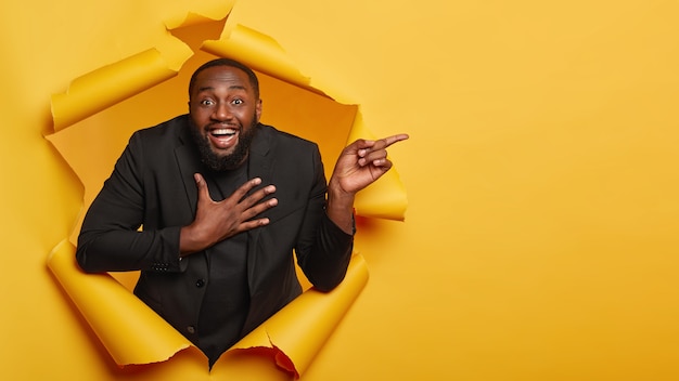 Positive unshaven businessman laughs pleasantly, keeps palm on chest, dressed elegantly, has friendly expression, points away on blank space, stands in hole of colored paper