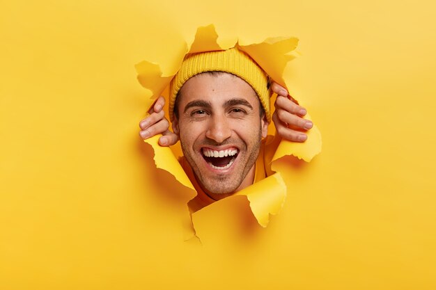 Positive unshaven male adult looks happily through yellow paper, shows only face