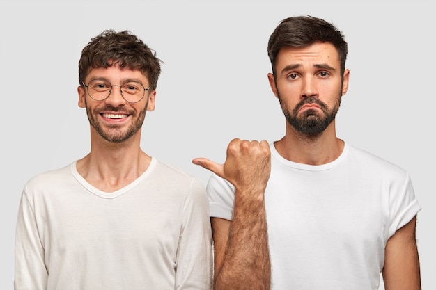 Positive unshaven young man has satisfied facial expression, being in good mood, bearded guy with discontent look points at friend