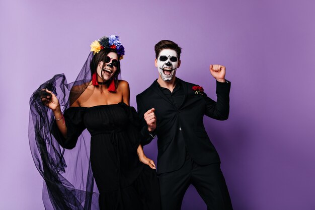 Positive vampires in black clothes dancing together. Smiling mexican muerte couple posing on purple background.