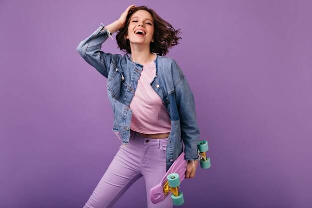 Positive white girl in pink pants posing with skateboard. Blithesome curly woman smiling.