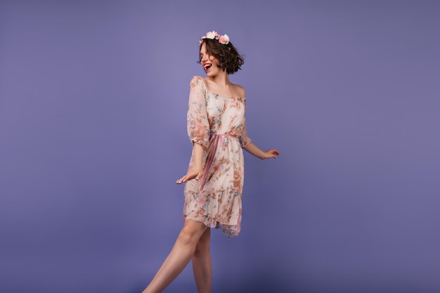 스튜디오에서 춤을 추는 세련된 여름 드레스를 입은 긍정적인 백인 여성 화환에서 사진 촬영을 즐기는 사랑스러운 곱슬 소녀