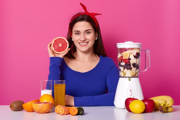 Positive woman in blue jumper and headband, prepares healthy juice, uses fresh ingredients, adds cut fruits in blender jar, holds slice of grapefruit likes smoothie in morning. Vegeterian food concept