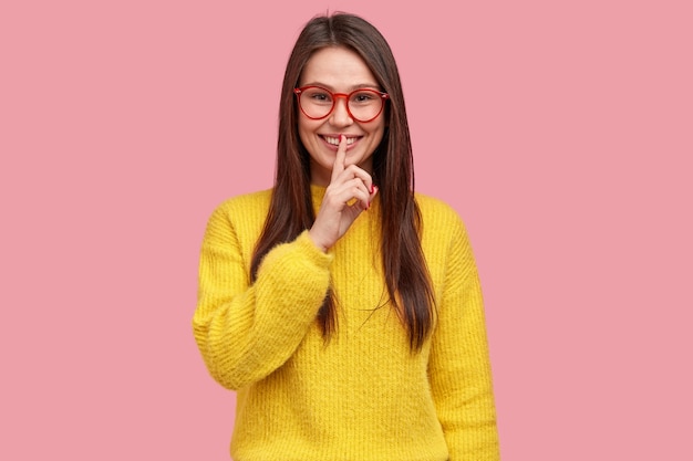 Positive woman demonstrates hush gesture, keeps fore finger over lips, dressed in casual outfit, gossips with best friend, tells secret information