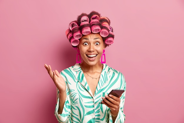 Позитивная женщина в шелковом костюме для сна использует загрузку мобильного телефона, новое приложение делает вьющиеся волосы с помощью бигуди, изолированных на розовой стене