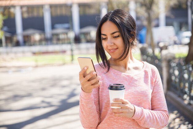 Positive woman drinking coffee and using smartphone outdoors