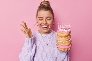 Позитивная женщина наслаждается празднованием дня рождения, держит поднятую ладонь, радостно хихикает, держит кучу вкусных пончиков со свечами, носит случайный джемпер, изолированный на розовом фоне. праздничный повод