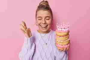 Бесплатное фото Позитивная женщина наслаждается празднованием дня рождения, держит поднятую ладонь, радостно хихикает, держит кучу вкусных пончиков со свечами, носит случайный джемпер, изолированный на розовом фоне. праздничный повод