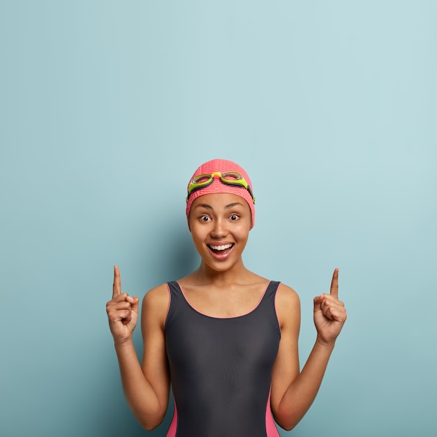 Positive woman enjoys water sport, dressed in black swimsuit, swimming hat and goggles, points above on free space, advertises accessories for diving, prepares for contest. Sport and promotion concept