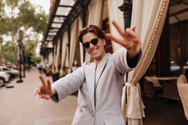 Positive woman in grey jacket and sunglasses showing peace signs outside Cheerful darkhaired girl in suit widely siles and walks around city