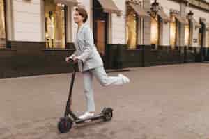 Free photo positive woman in grey suit riding scooter outdoors. charming short-haired girl in jacket and pants smiling and walking around city