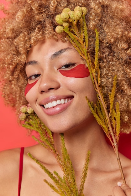 Positive woman has toothy smile on face holds plants near face wears red hydrogel patches under eyes to remove wrinkles uses natural cosmetic products for skin moisturizing. facial skin care concept