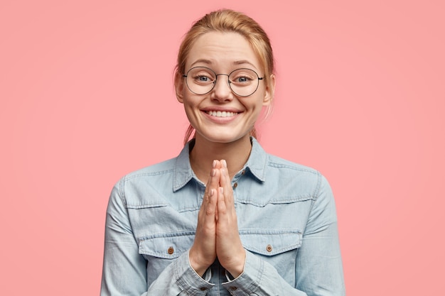 Positive woman in jean jacket, presses palms together in buddhist gesture, bows to greet friend from abroad, prays for something good, isolated over pink wall