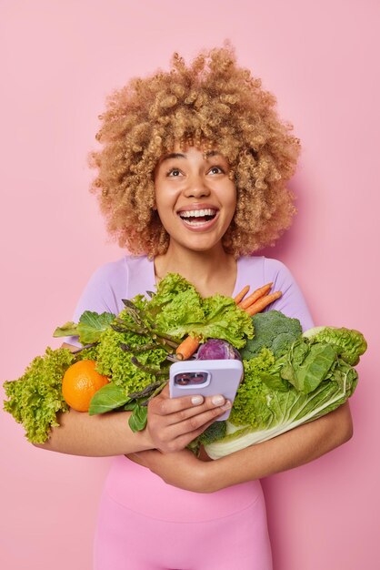 Positive woman looks happily above uses mobile phone for home delivery service carries fresh green vegetables makes order online eats healthy food isolated over pink background Grocery concept