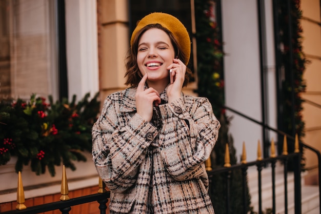 주황색 베레모와 코트에 긍정적 인 여성이 도시 벽에 전화로 이야기하는 동안 웃음