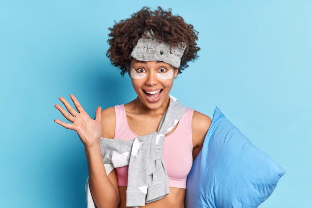 Positive woman raises palm glad to have rest ater hard week enjoys laziness and relaxation keeps palm raised holds pillow isolated over blue wall excited with good dreams